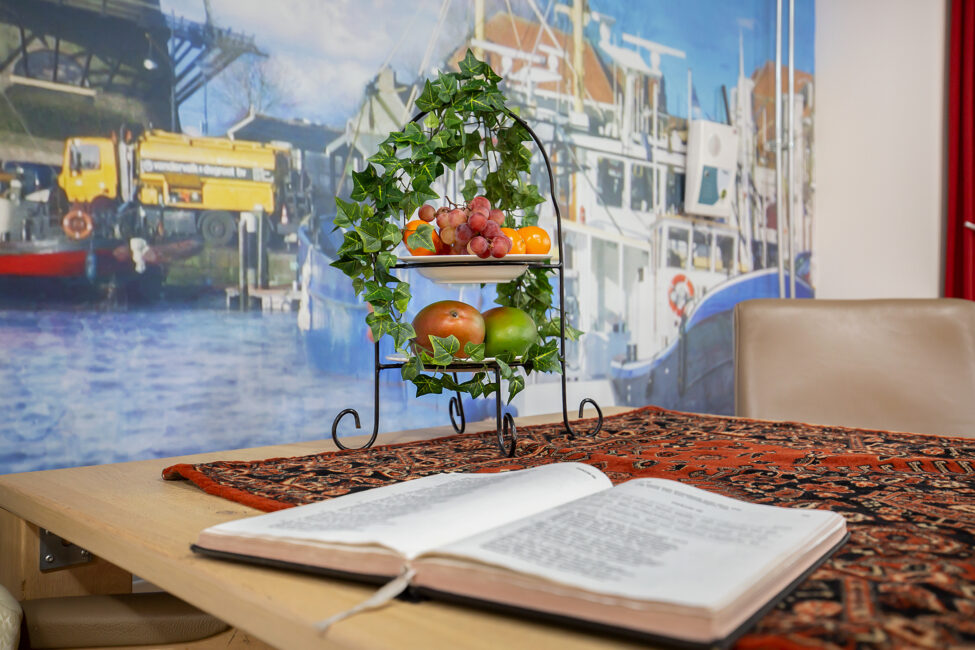 Open bijbel op tafel met fruitschaal op een kleurrijk kleed, tegen een achtergrond van een haven met boten en gebouwen.