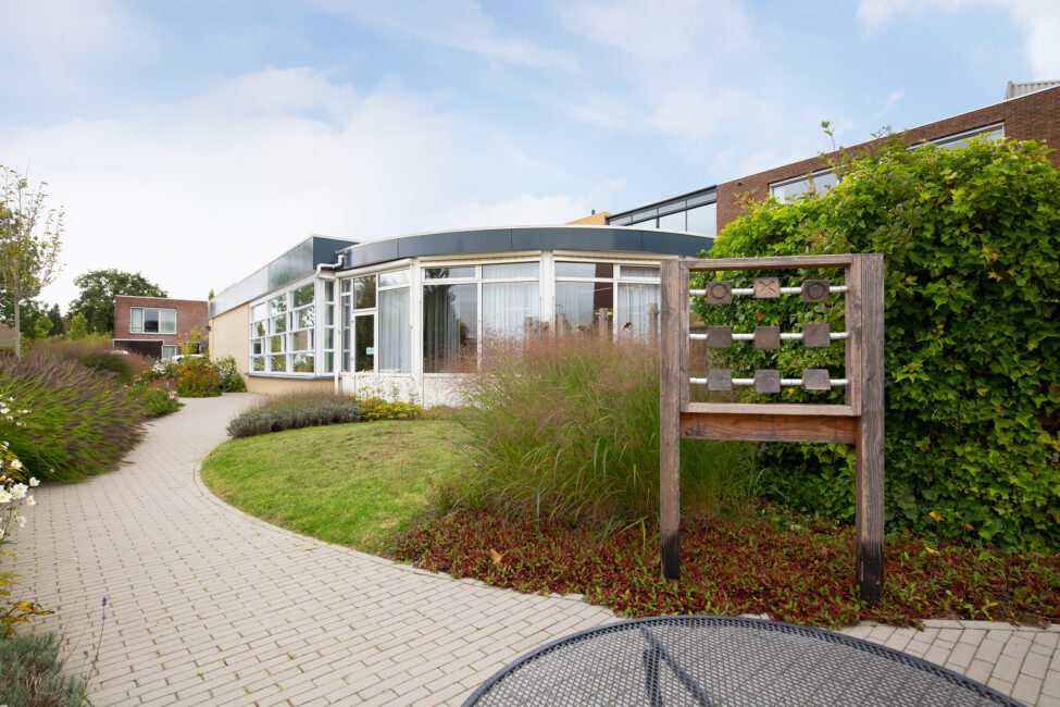 Moderne zorginstelling met glazen gevel, omgeven door een tuin met een kronkelend pad en groene struiken onder een blauwe hemel.
