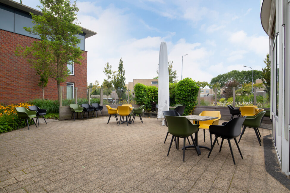 Buitenterras met tafels en stoelen, omgeven door groene struiken en bomen, naast een modern gebouw onder een blauwe hemel.