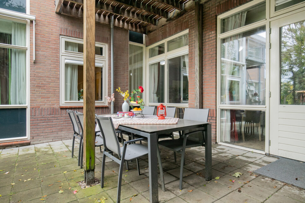 Terras met een tafel en vier stoelen, omringd door bakstenen muren en ramen. Op tafel staan bloemen en decoraties.