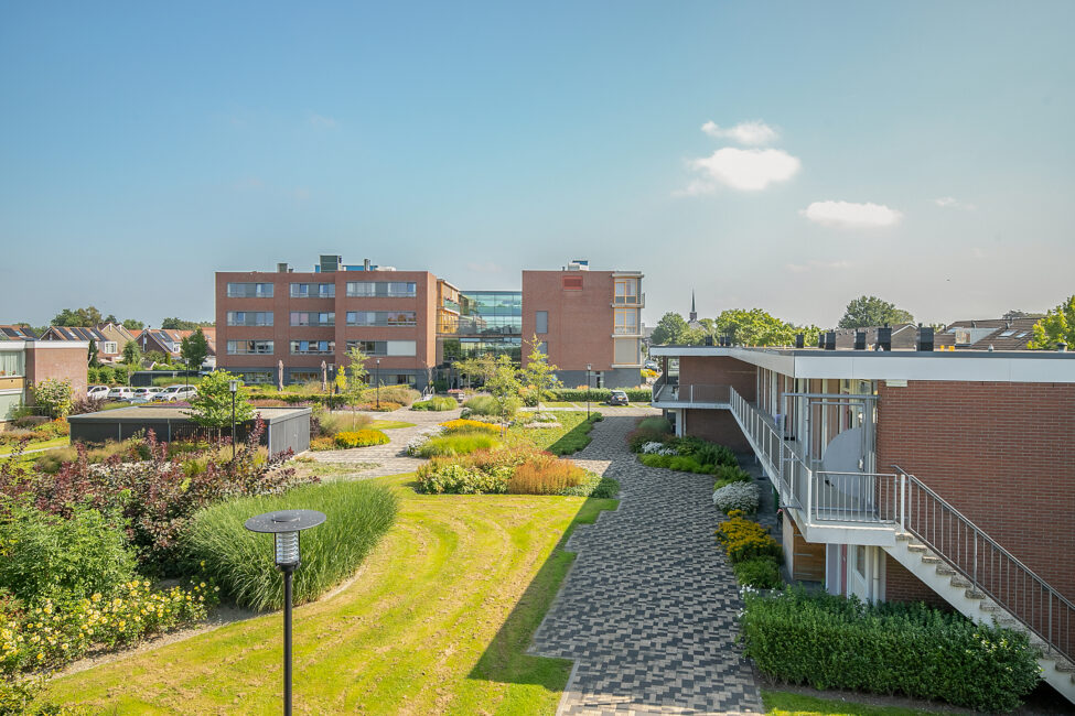 Luchtfoto van een modern zorgcentrum met bakstenen gebouwen, groene tuinen en een geplaveid pad onder een heldere blauwe hemel.