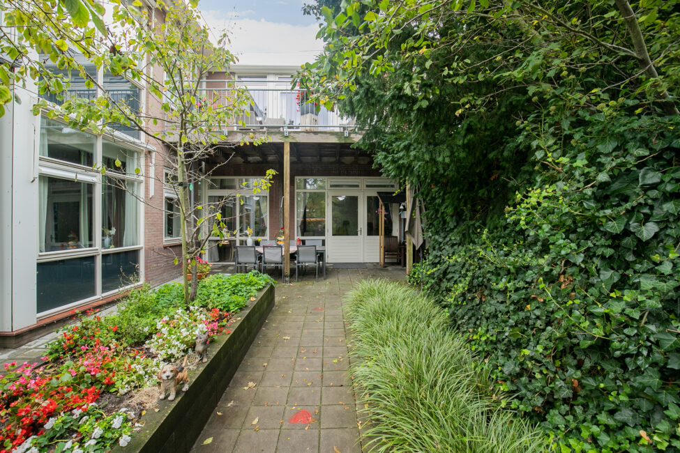 Tuinpad met bloembedden en groene struiken leidt naar een overdekt terras met tafels en stoelen bij een gebouw.