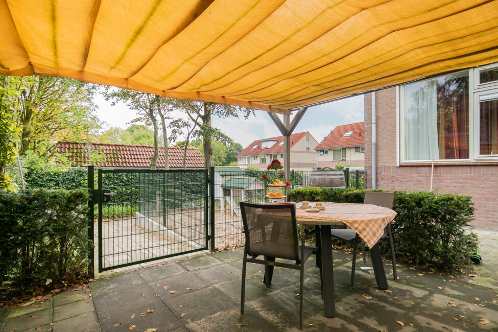 Tuin met een tafel en twee stoelen onder een oranje zonnescherm. Op de tafel staat een vaas met bloemen. Hek en huizen op de achtergrond.