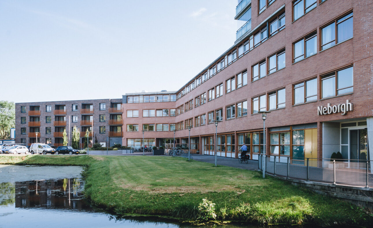 Afbeelding van een modern bakstenen gebouw met meerdere verdiepingen, omgeven door een grasveld en een vijver op de voorgrond.