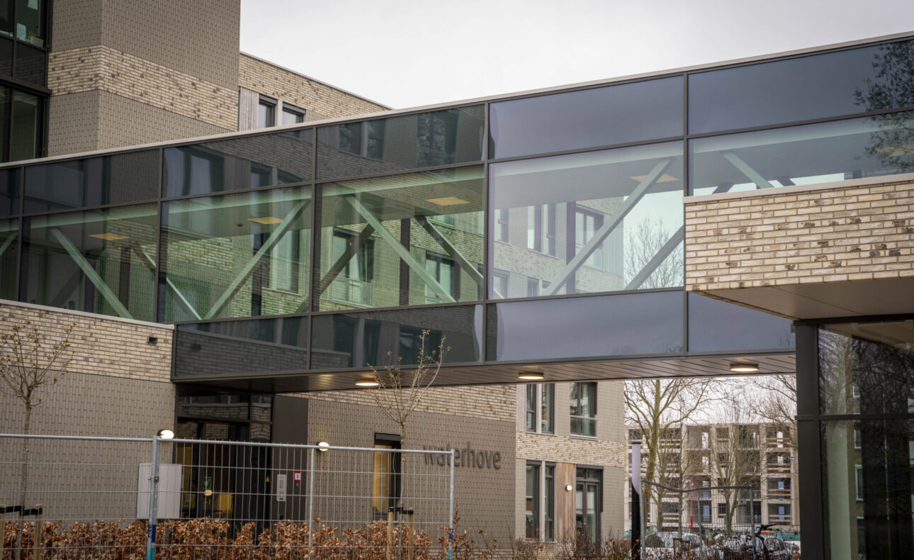 Moderne architectuur van een gebouw met glazen loopbrug en bakstenen gevel, omgeven door een hek en kale bomen.