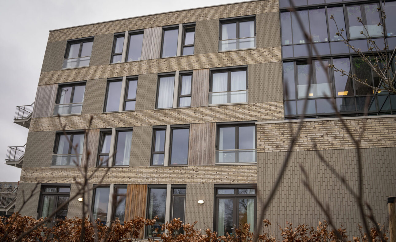 Buitenaanzicht van een modern, vier verdiepingen hoog gebouw met bakstenen gevel en grote ramen, omgeven door kale takken.