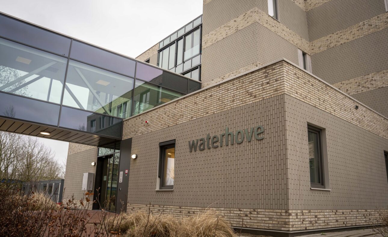 Buitenaanzicht van een modern gebouw met de naam "Waterhove" op de gevel, met glazen loopbrug en bakstenen muren.