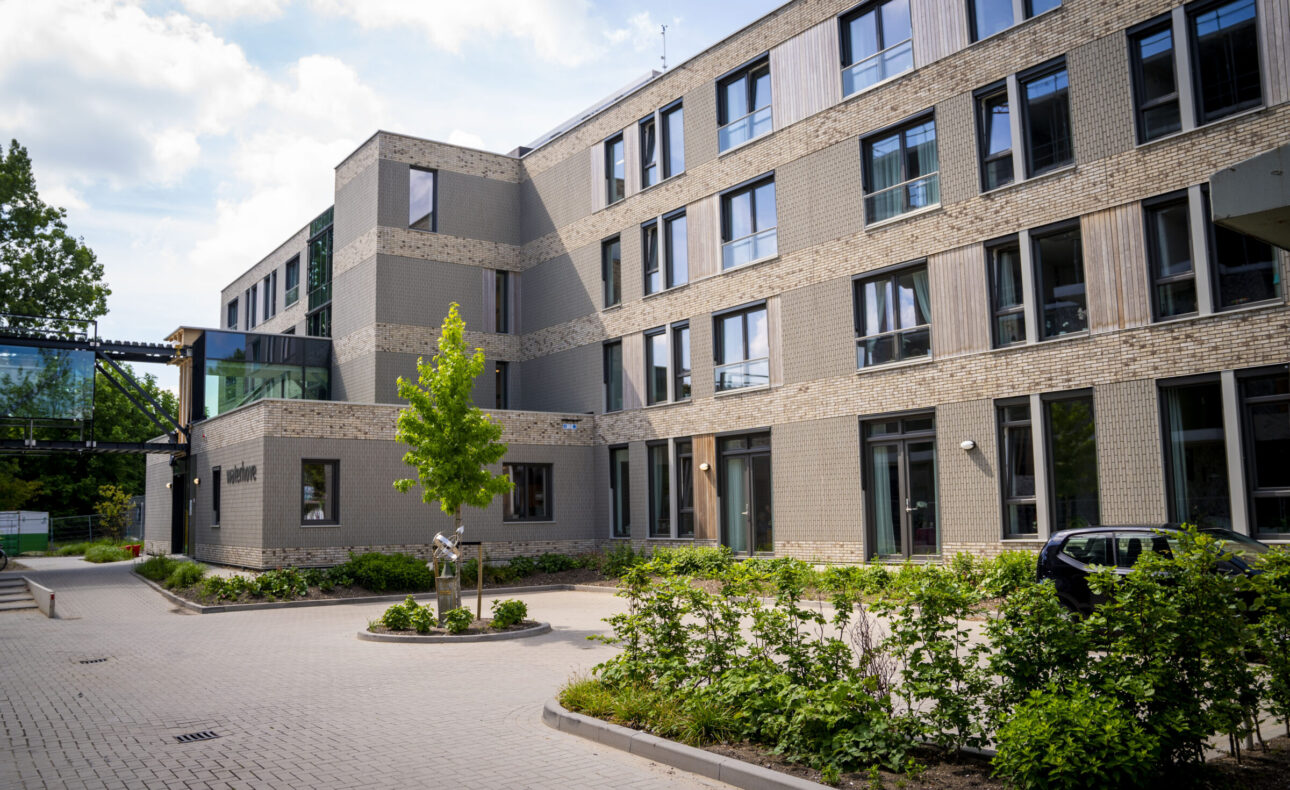 Buitenaanzicht van een modern gebouw met meerdere verdiepingen, omgeven door groen en een betegeld pad onder een bewolkte hemel.