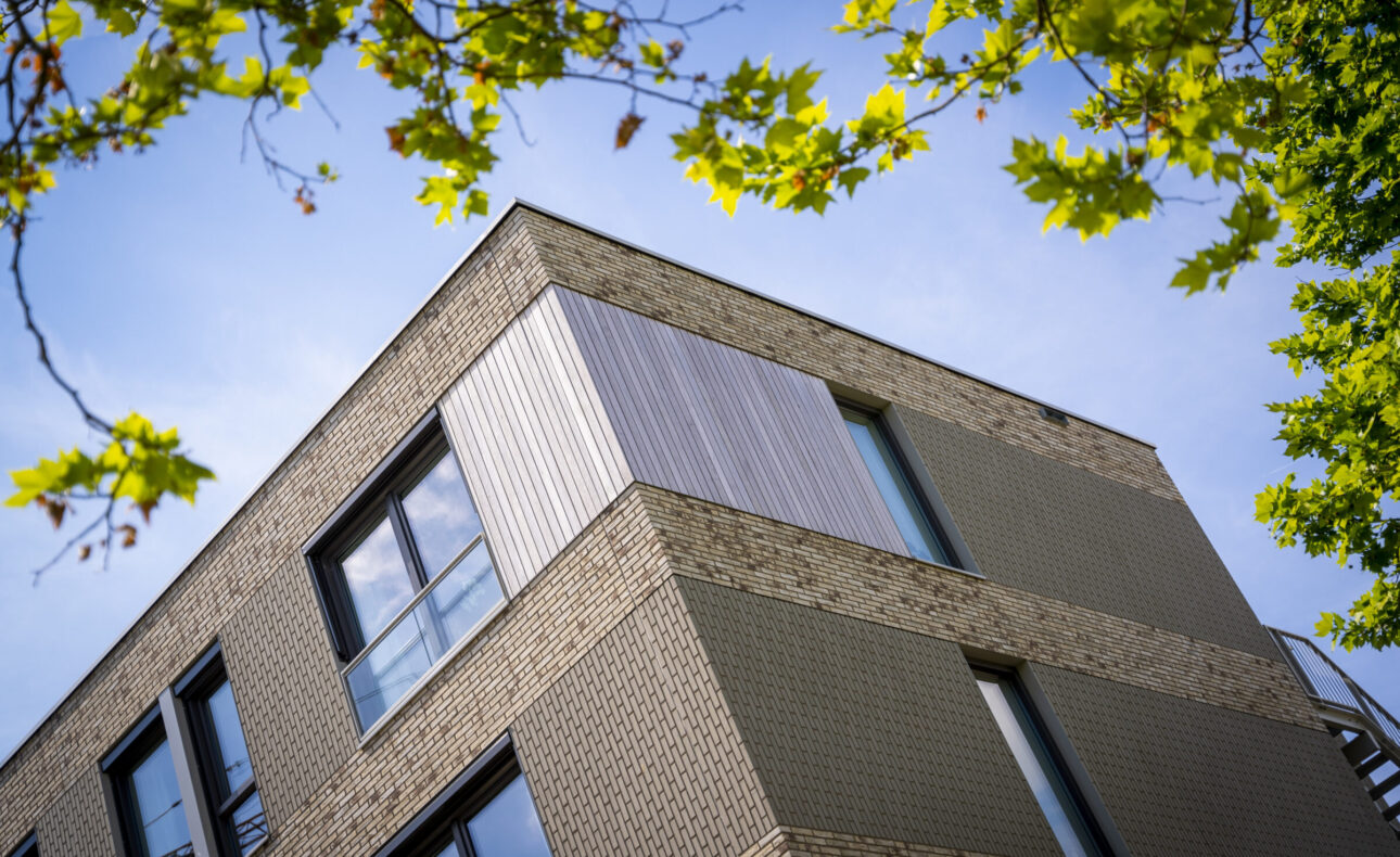 Bovenaanzicht van een modern gebouw met bakstenen en metalen gevel, omringd door groene bladeren onder een blauwe hemel.