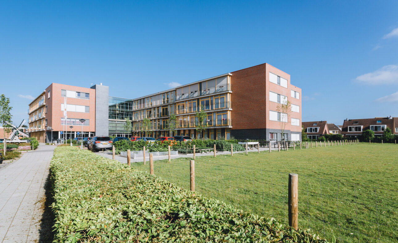 Buitenaanzicht van een modern gebouw met rode bakstenen en grote ramen, omgeven door een groen grasveld en een haag.