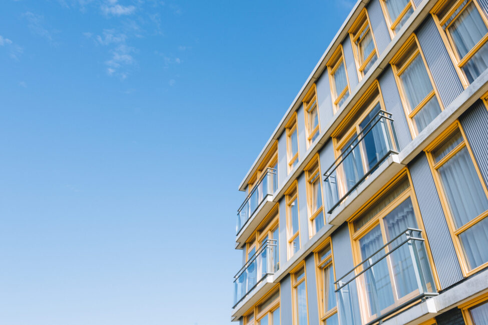 Buitenkant van een modern gebouw met meerdere verdiepingen, grote ramen en balkons, tegen een heldere blauwe lucht.