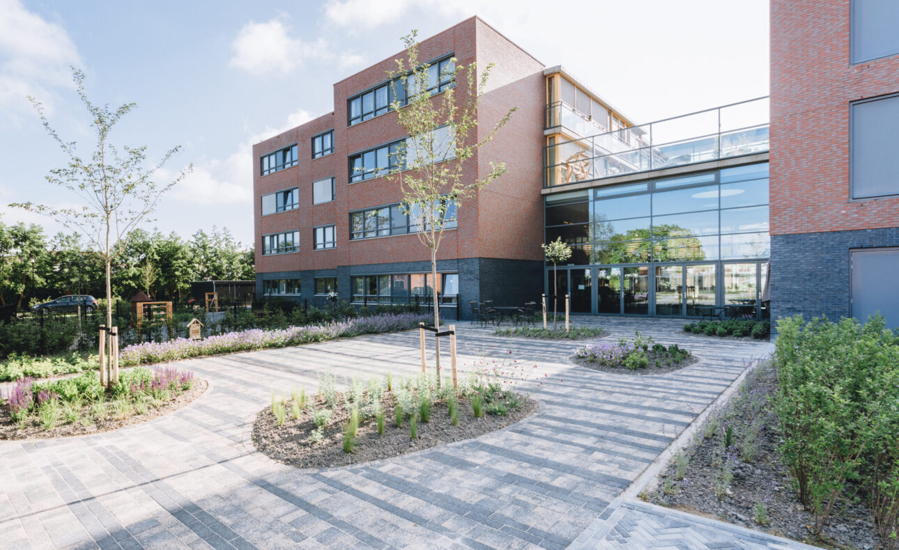 Moderne bakstenen gebouw met grote ramen, omgeven door een tuin met jonge bomen en planten, onder een blauwe lucht.