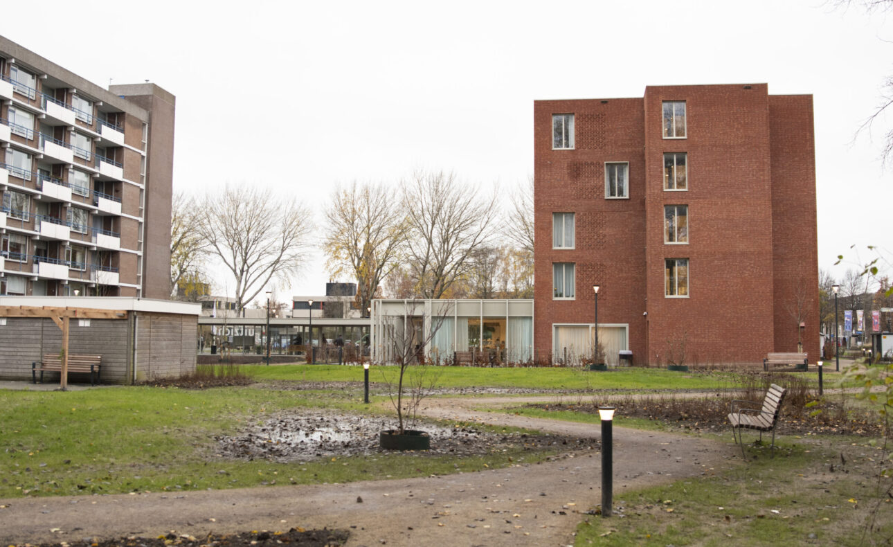 Appartementengebouw met rode bakstenen en een tuin met wandelpaden en bankjes, omgeven door bomen en een ander gebouw.