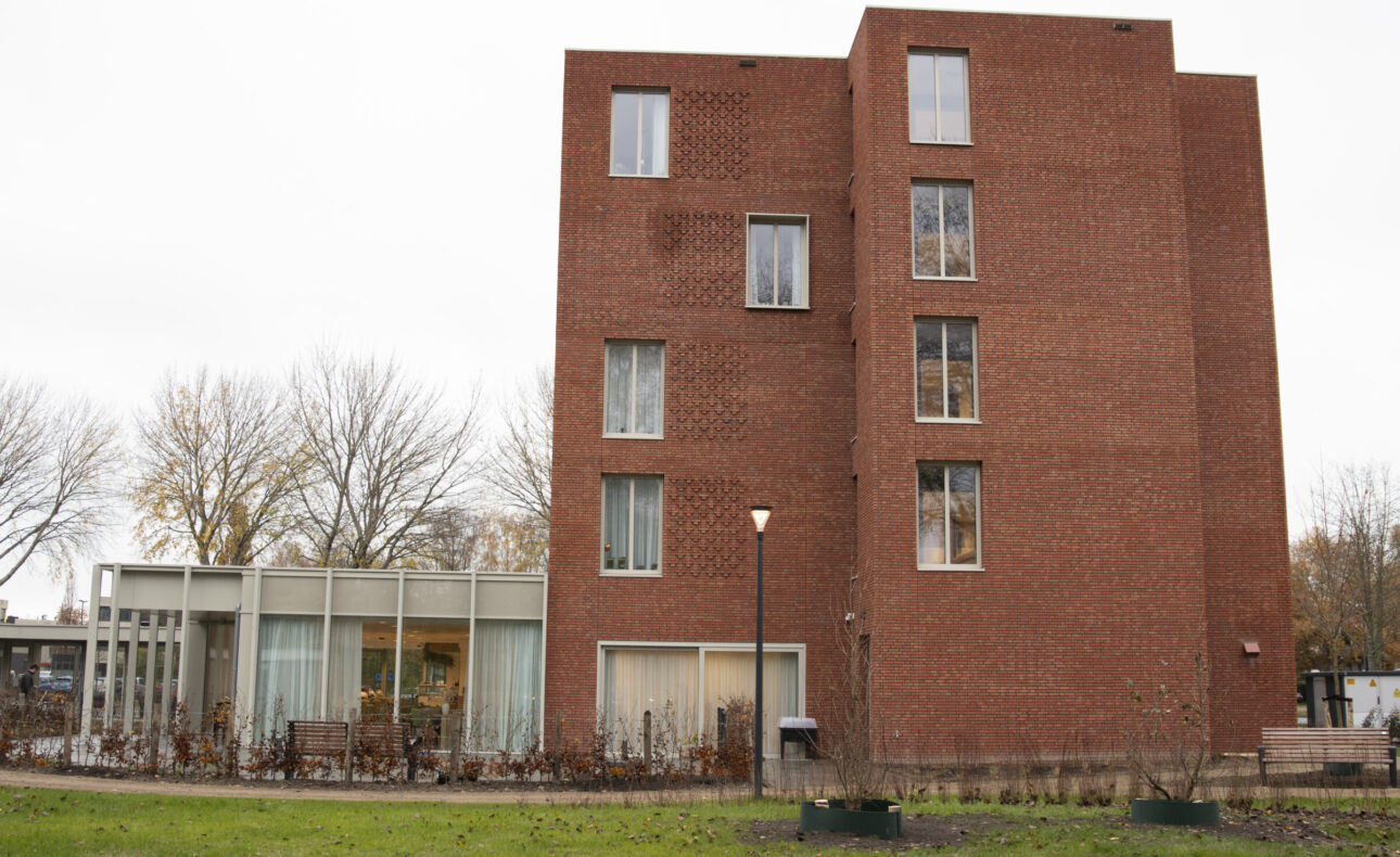 Afbeelding van een bakstenen gebouw met meerdere verdiepingen, omgeven door een grasveld en kale bomen op de achtergrond.
