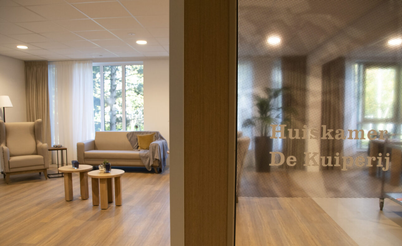 Een gezellige woonkamer met comfortabele stoelen, een bank en tafels. Op de deur staat "Huiskamer De Kuiperij".