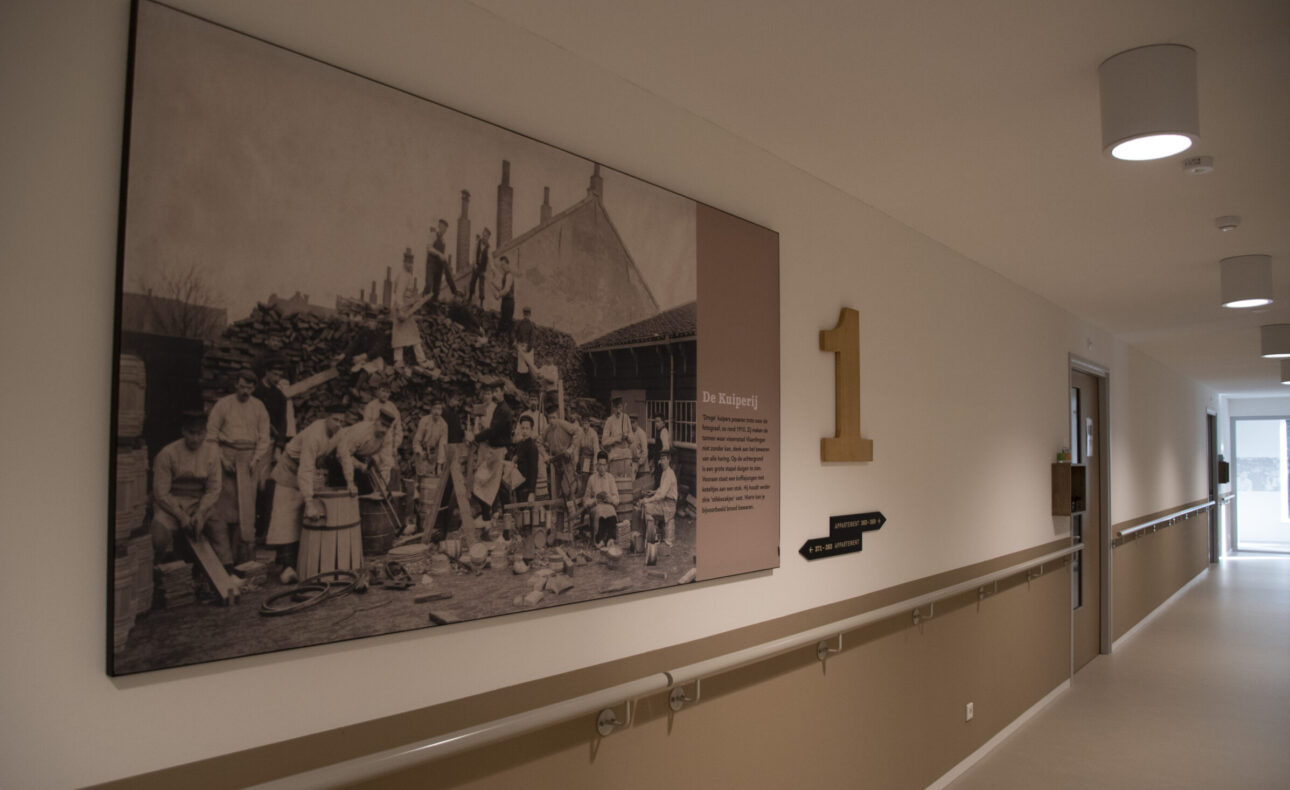 Zwart-witfoto van een groep mensen in een gang van een gebouw, met een groot nummer één op de muur ernaast.