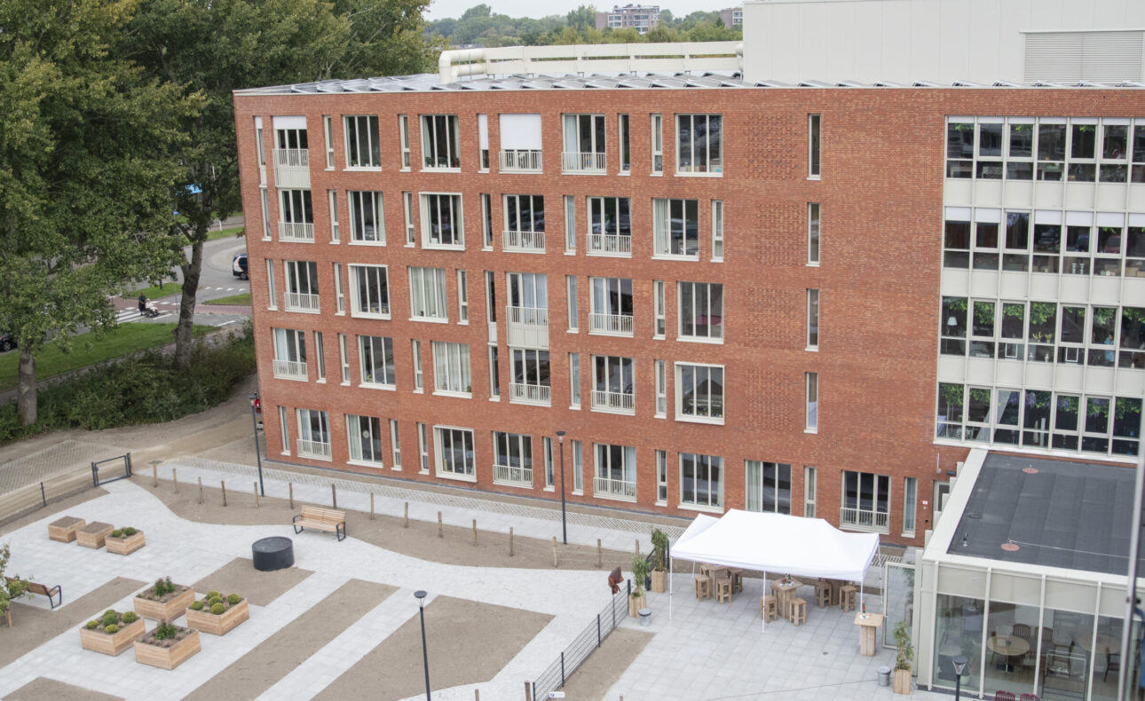 Luchtfoto van een modern bakstenen gebouw met grote ramen, omgeven door een plein met bomen en zitplaatsen.
