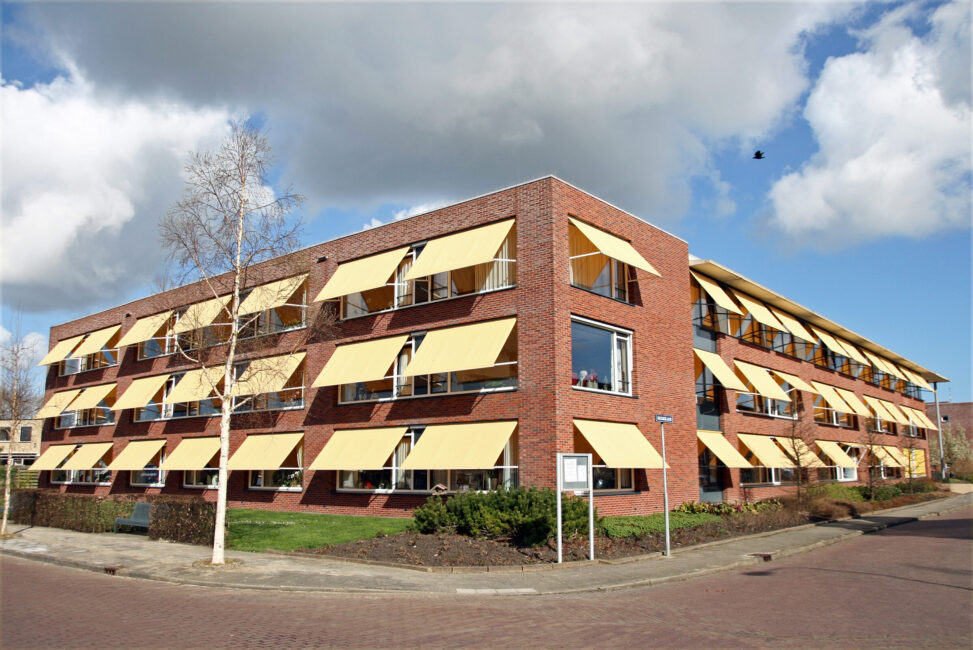Een bakstenen gebouw met drie verdiepingen en gele zonneschermen voor de ramen, onder een blauwe lucht met wolken.