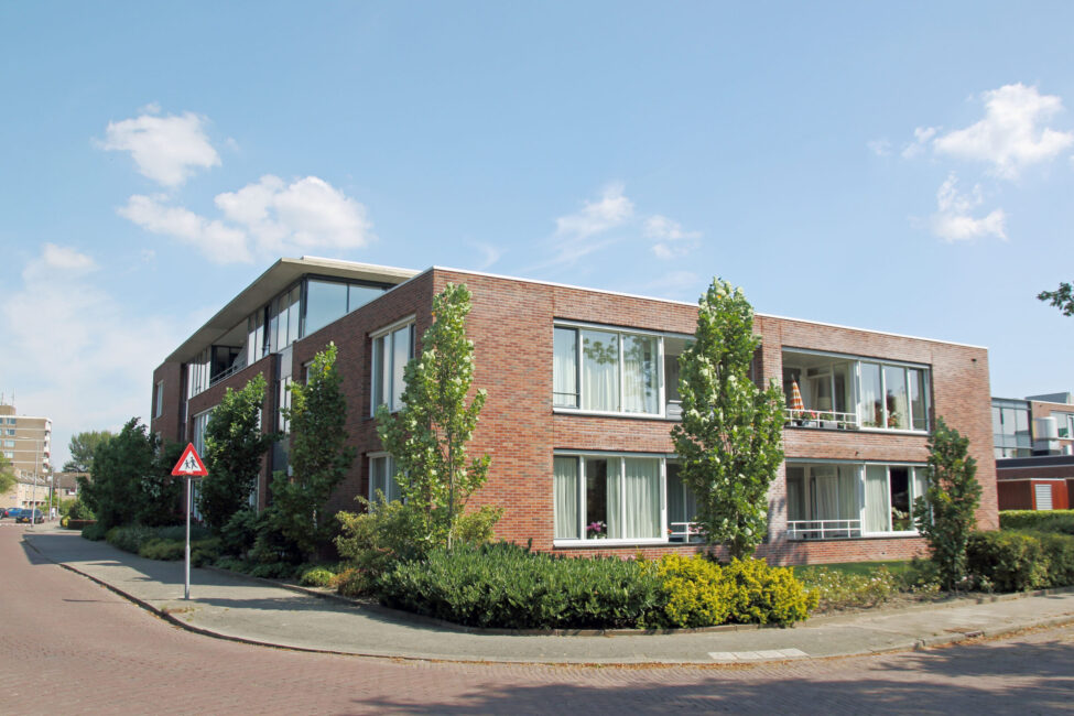 Een modern bakstenen gebouw met grote ramen en groene struiken eromheen, gelegen aan een rustige straat op een zonnige dag.