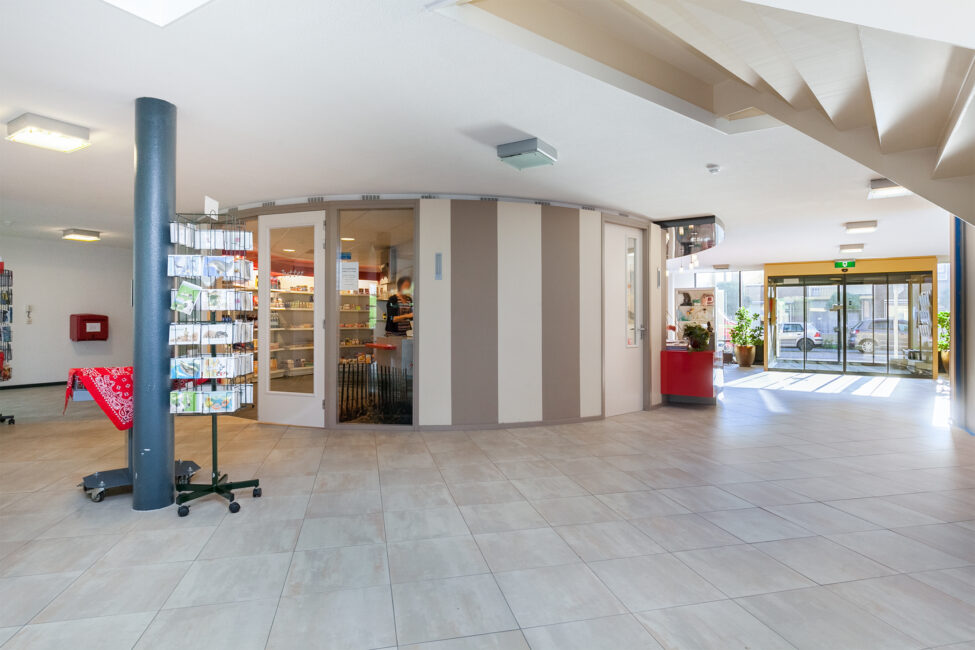Een lichte, ruime hal met een ronde kiosk in het midden, een rek met kaarten links en een glazen deur naar buiten rechts.