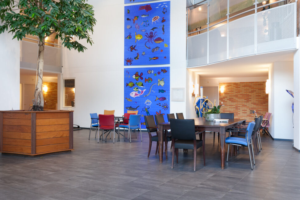 Een moderne gemeenschappelijke ruimte met een grote blauwe muurschildering, diverse stoelen en een grote plant in een houten bak.