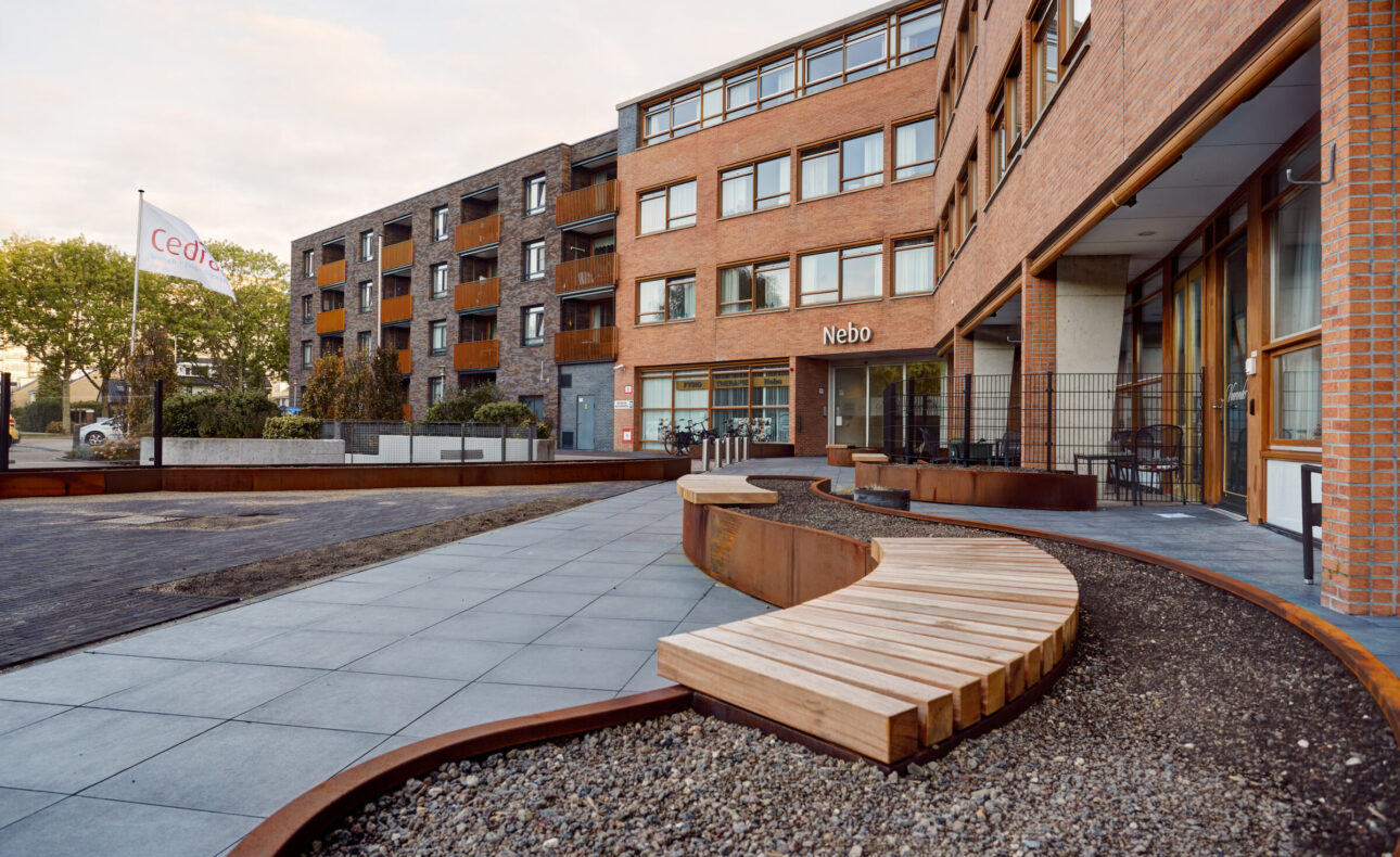 Buitenaanzicht van een modern gebouw met bakstenen gevel en grote ramen, omgeven door een betegeld pad en houten bankjes.