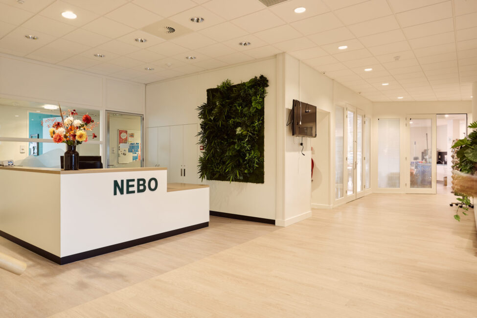 Ingang van een modern zorgcentrum met een receptiebalie, bloemen, een groene plantenwand en glazen deuren in een lichte ruimte.