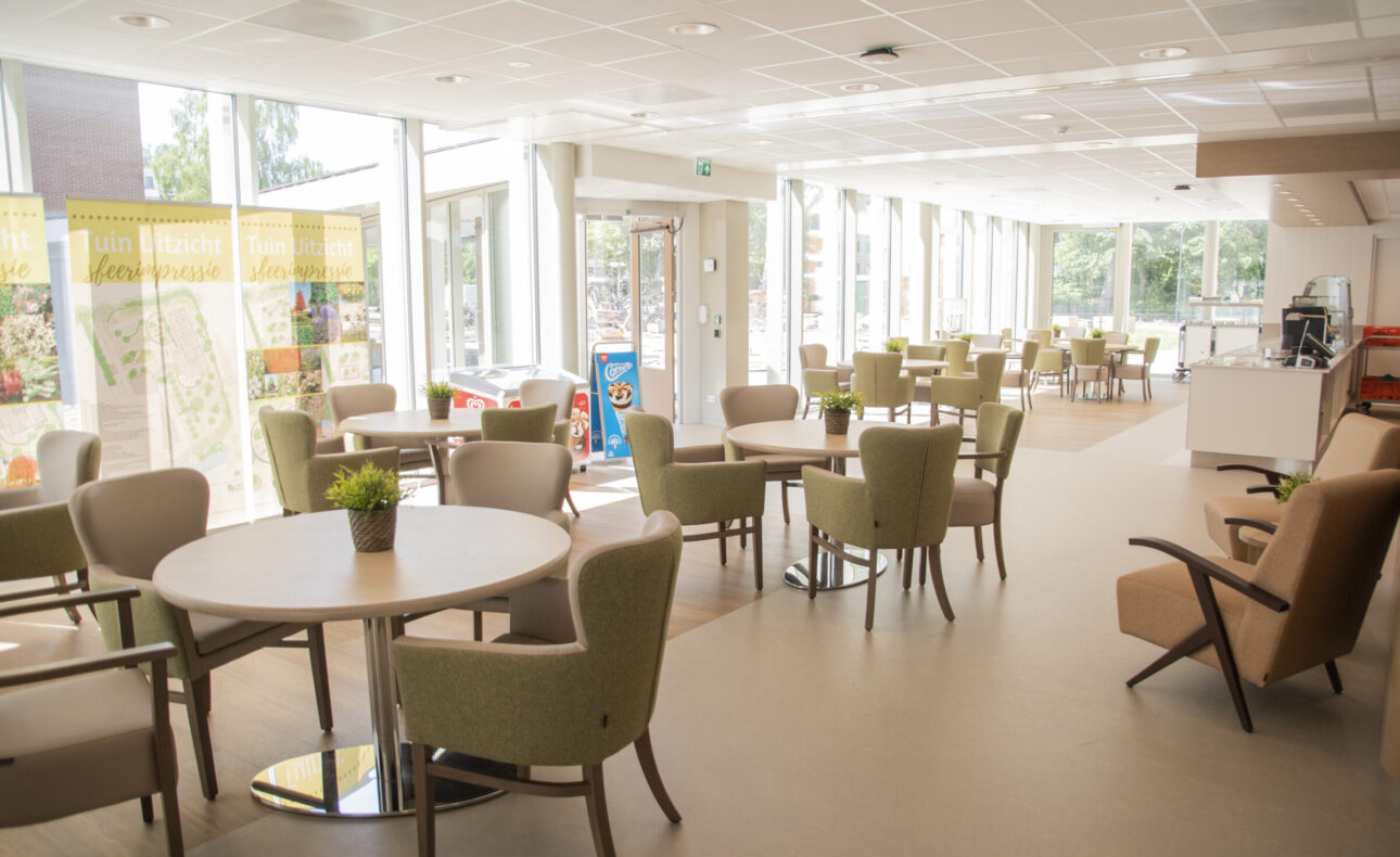 Een lichte, moderne gemeenschappelijke ruimte met tafels en stoelen, grote ramen en uitzicht op een groene buitenomgeving.