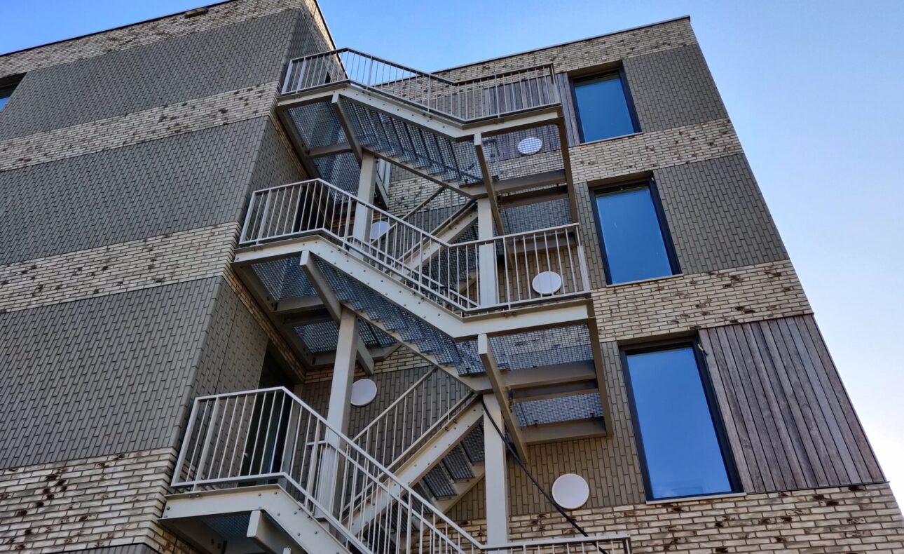 Buitentrap van een modern gebouw met bakstenen en grote ramen, gefotografeerd vanuit een laag standpunt tegen een blauwe lucht.