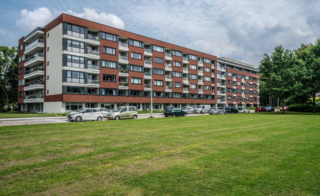 Afbeelding van een modern, rood-bruin appartementencomplex met meerdere verdiepingen, omgeven door een grasveld en bomen.
