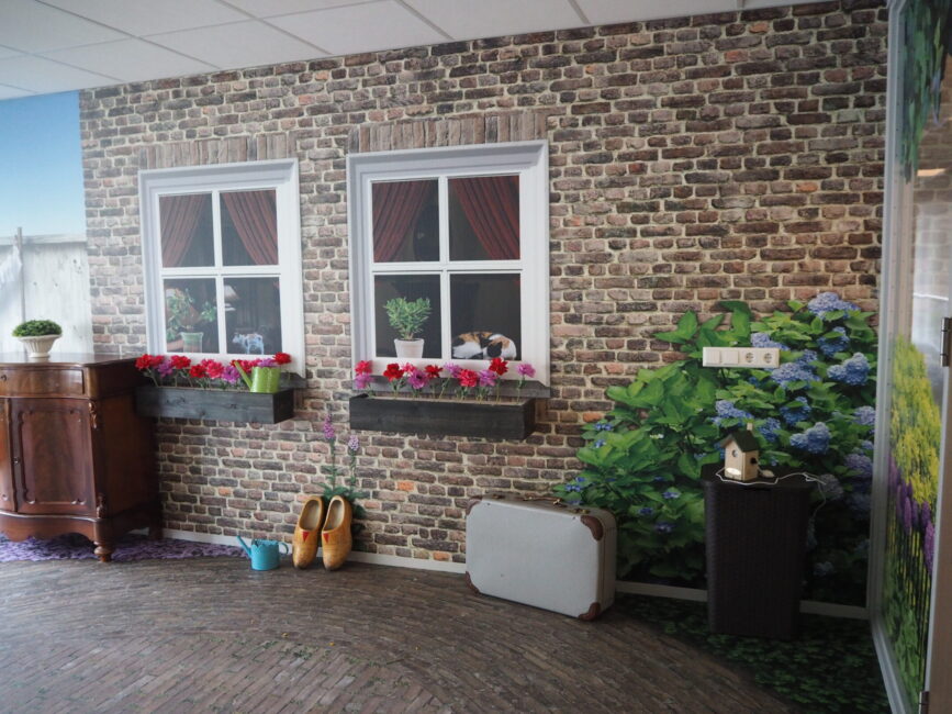 Een kamer met een muur beschilderd als bakstenen gevel met ramen, bloembakken en klompen. Er staat een houten kast links.