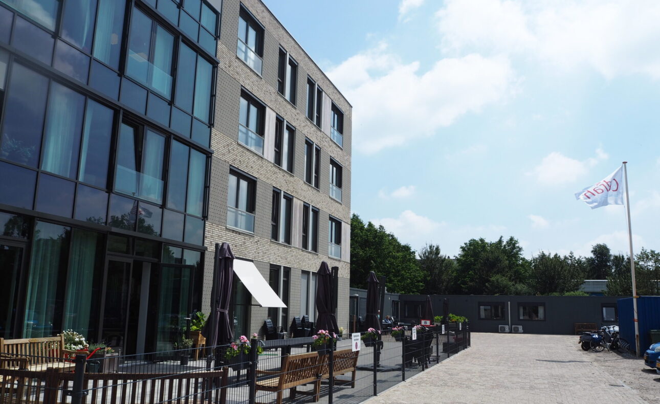 Modern gebouw met glazen gevel en meerdere verdiepingen. Voor het gebouw staan houten banken en bloembakken op een terras.