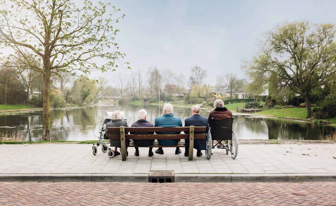 Vijf ouderen zitten op een bankje en in een rolstoel, kijkend naar een rustige vijver met bomen op de achtergrond.