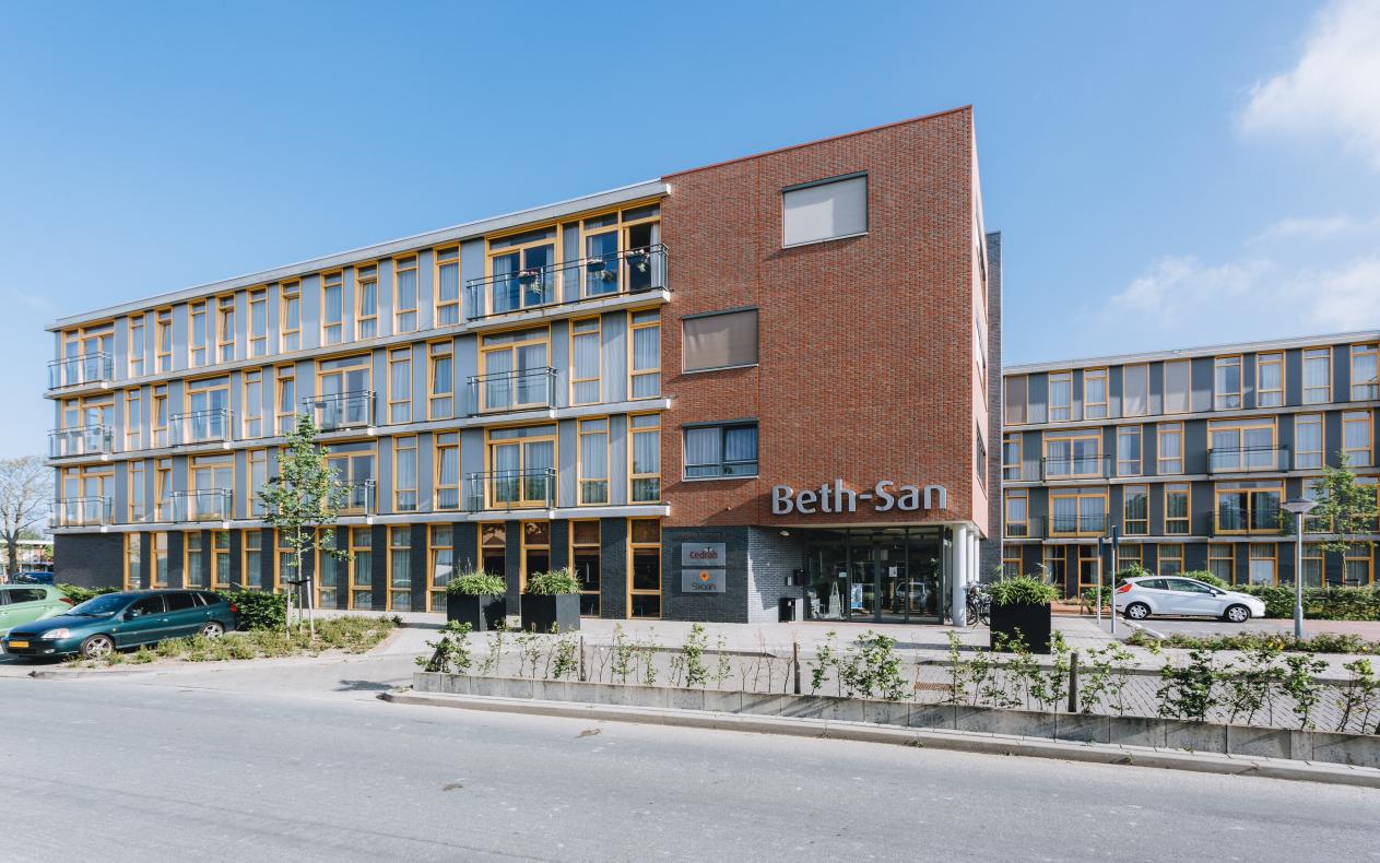 Afbeelding van een modern gebouw met drie verdiepingen, grote ramen en een rode bakstenen gevel. Er staat "Beth-San" op de gevel.
