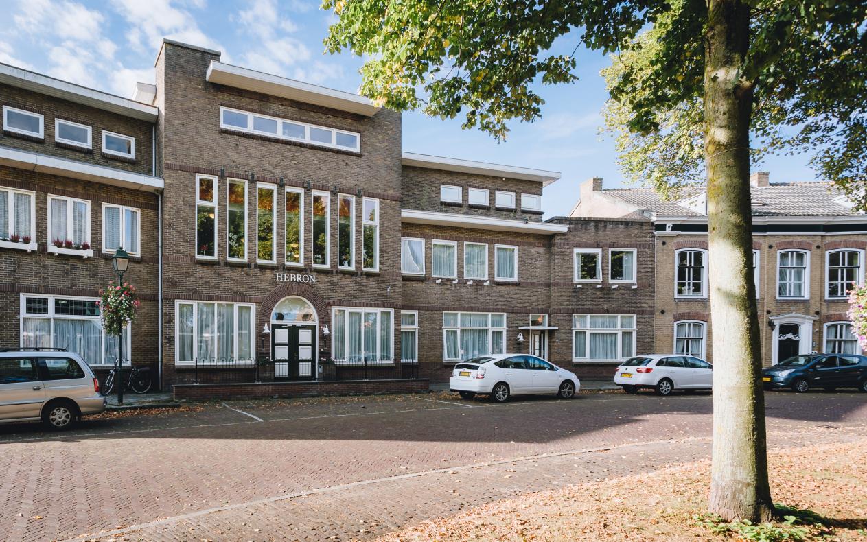 Een straatbeeld met een rij bakstenen gebouwen, geparkeerde auto's en een boom aan de rechterkant onder een blauwe hemel.