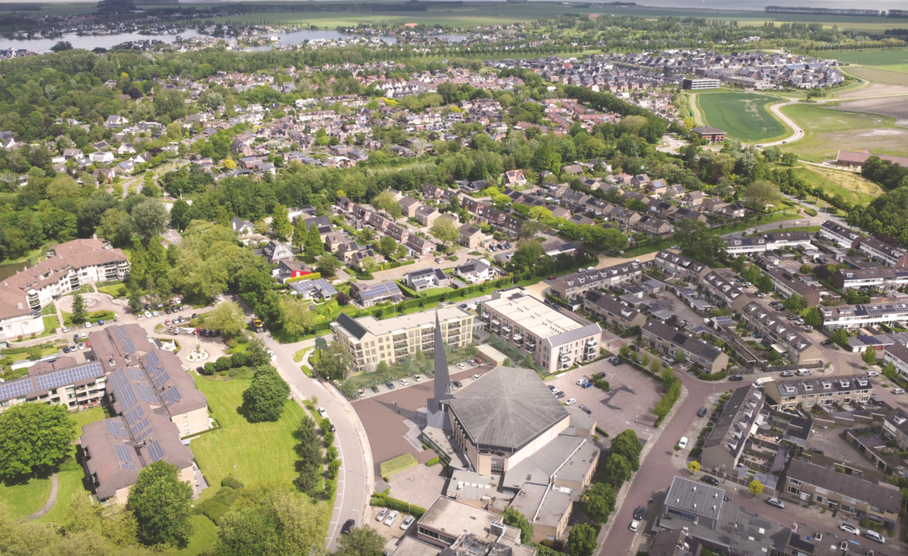 Luchtfoto van een woonwijk met huizen, wegen en groene gebieden, omringd door bomen en een rivier in de verte.