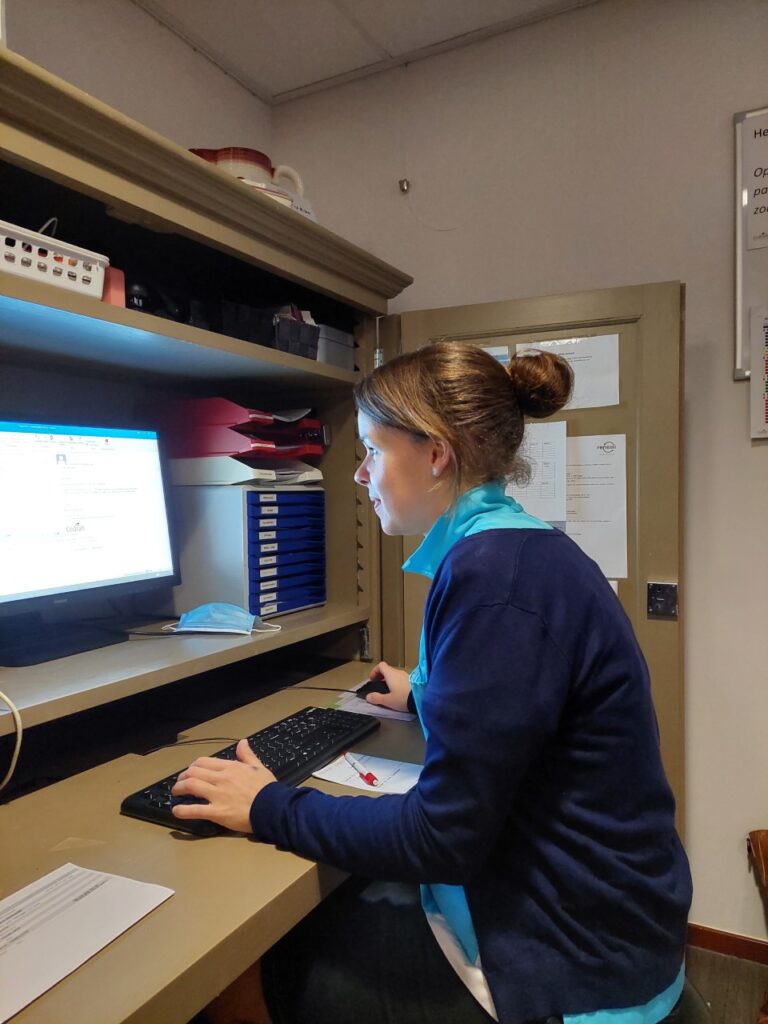 Vrouw werkt geconcentreerd op een computer in een kantooromgeving, zittend aan een bureau met documenten en mappen.