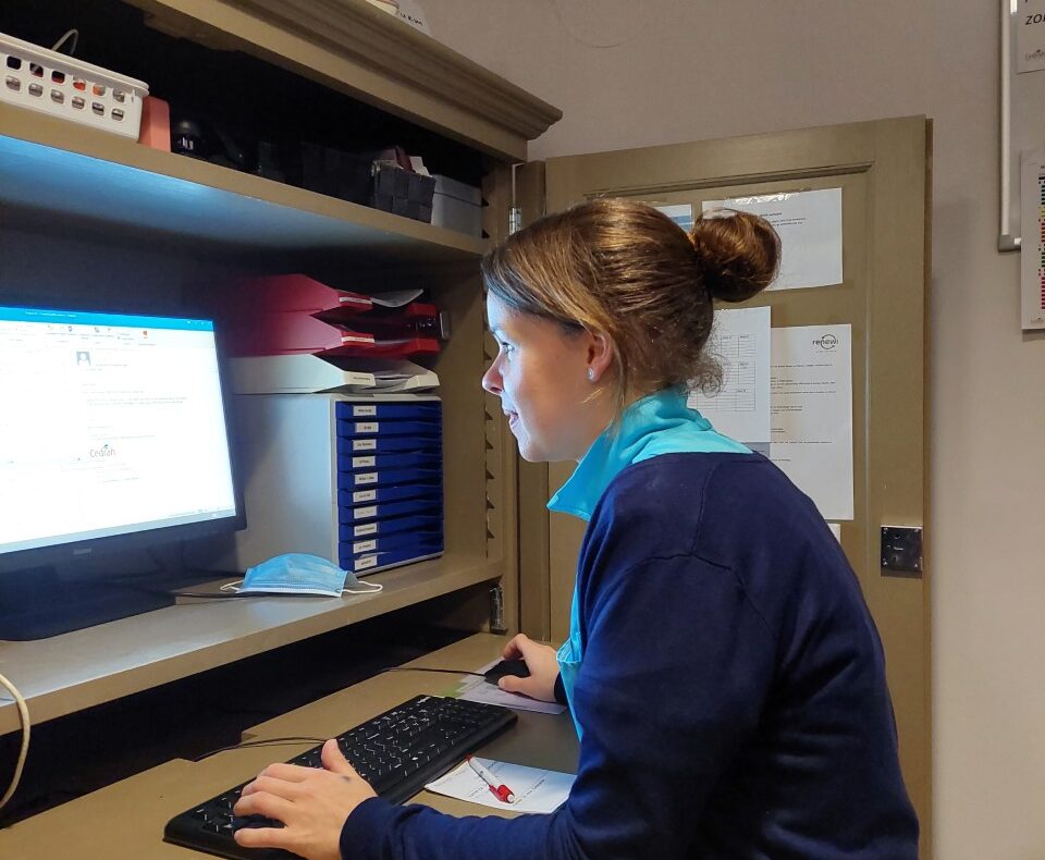 Vrouw werkt geconcentreerd op een computer in een kantooromgeving, zittend aan een bureau met documenten en mappen.