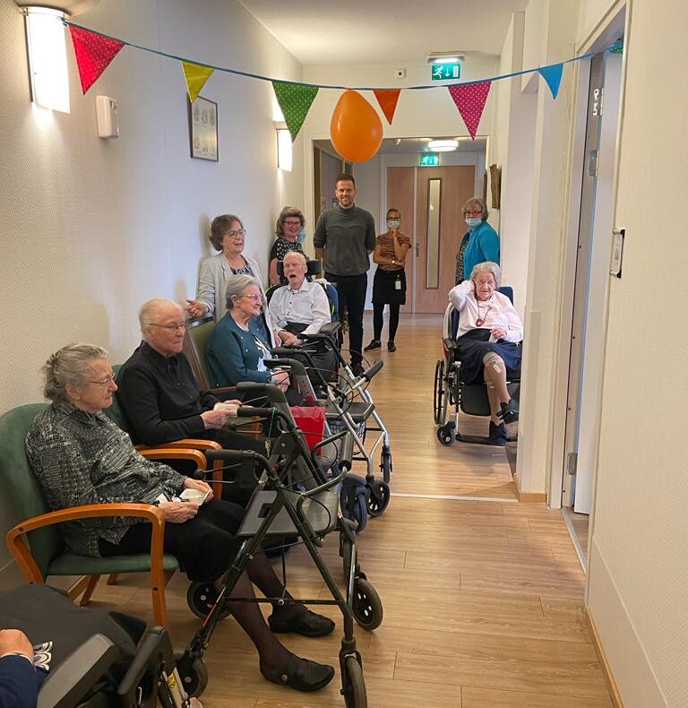 Groep ouderen in rolstoelen en met rollators in een gang, versierd met kleurrijke vlaggetjes, begeleid door verzorgers.