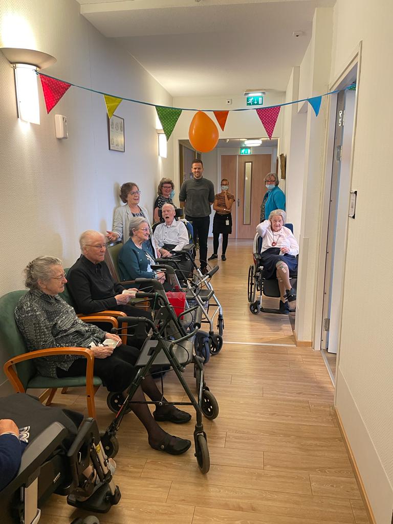 Groep ouderen in rolstoelen en met rollators in een gang, versierd met kleurrijke vlaggetjes, begeleid door verzorgers.