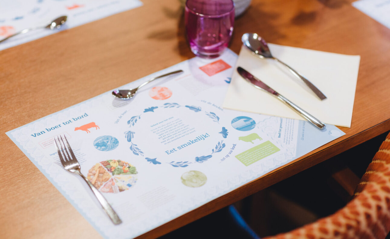 Een gedekte tafel met een placemat, bestek, een roze glas en een servet. De placemat toont illustraties en tekst.