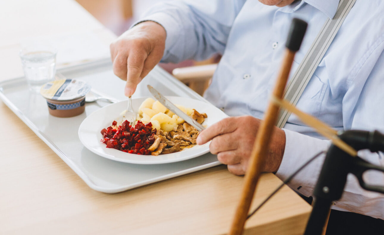 Een oudere man eet een maaltijd met aardappelen, rode bessen en vlees. Hij zit aan een tafel met een looprek naast zich.