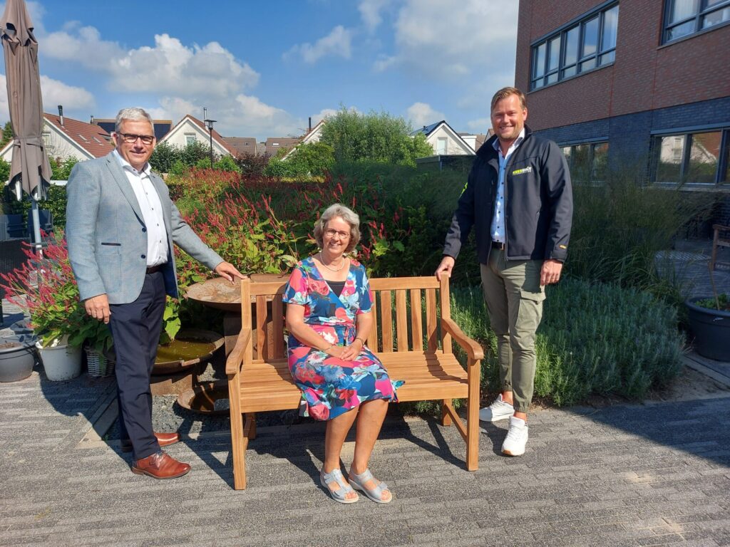 Drie mensen poseren bij een houten tuinbank op een zonnig terras, omringd door planten en een gebouw op de achtergrond.