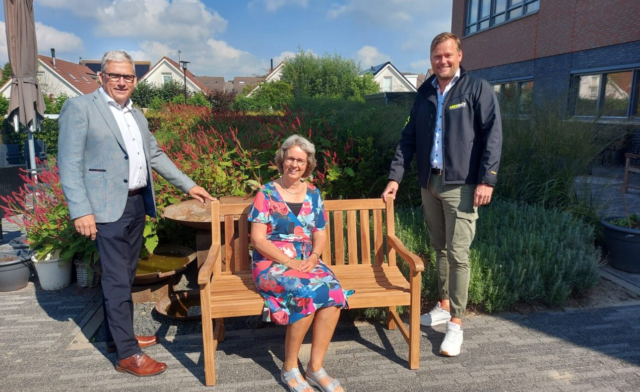 Drie mensen poseren bij een houten tuinbank op een zonnig terras, omringd door planten en een gebouw op de achtergrond.
