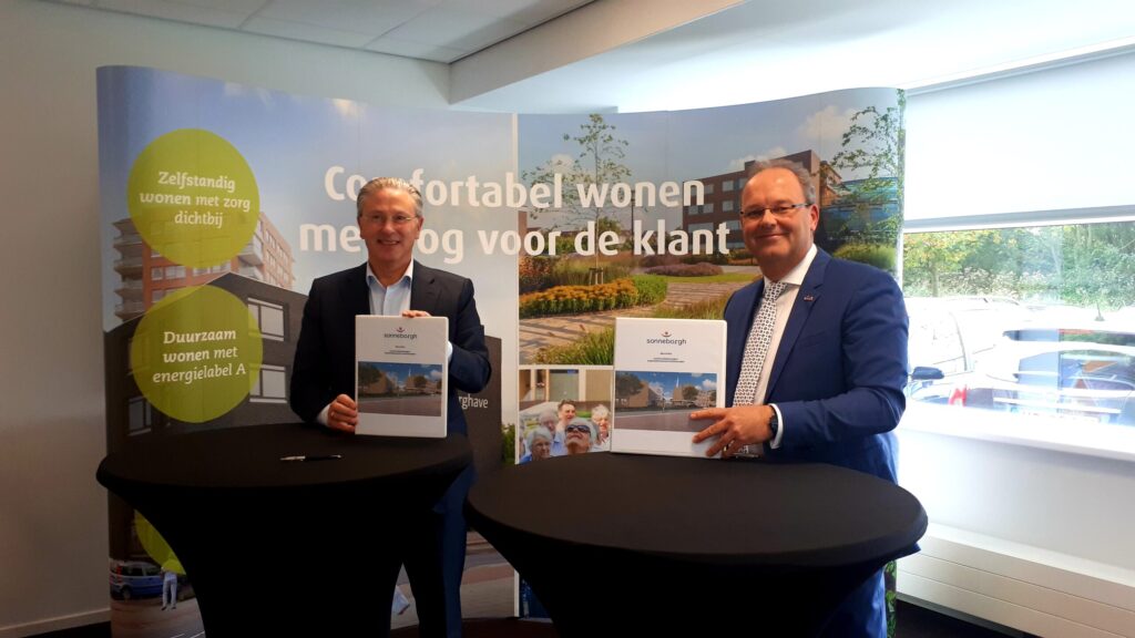 Twee mannen in pakken houden brochures vast bij een presentatie over comfortabel wonen, met banners op de achtergrond.
