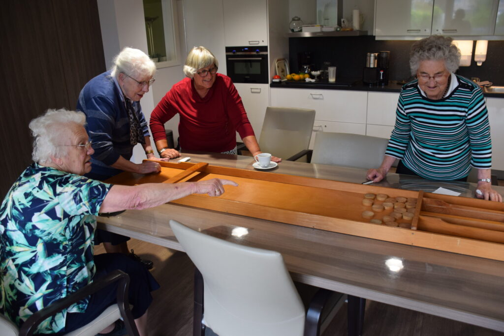 Vier oudere vrouwen spelen sjoelen aan een tafel in een moderne keuken. Ze lijken plezier te hebben tijdens het spel.