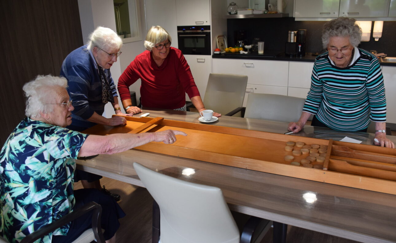 Vier oudere vrouwen spelen sjoelen aan een tafel in een moderne keuken. Ze lijken plezier te hebben tijdens het spel.
