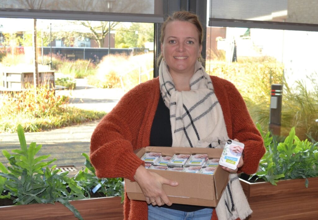 Vrouw met een oranje vest en sjaal houdt een doos met voedselproducten vast in een lichte ruimte met planten op de achtergrond.