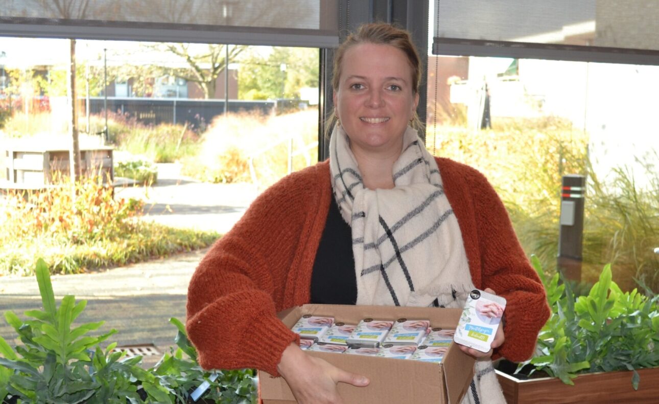 Vrouw met een oranje vest en sjaal houdt een doos met voedselproducten vast in een lichte ruimte met planten op de achtergrond.