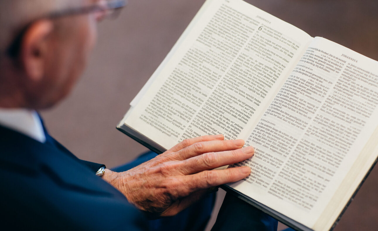 Een oudere man leest aandachtig in een open boek. Hij draagt een donkerblauw pak en zijn handen rusten op de pagina's.